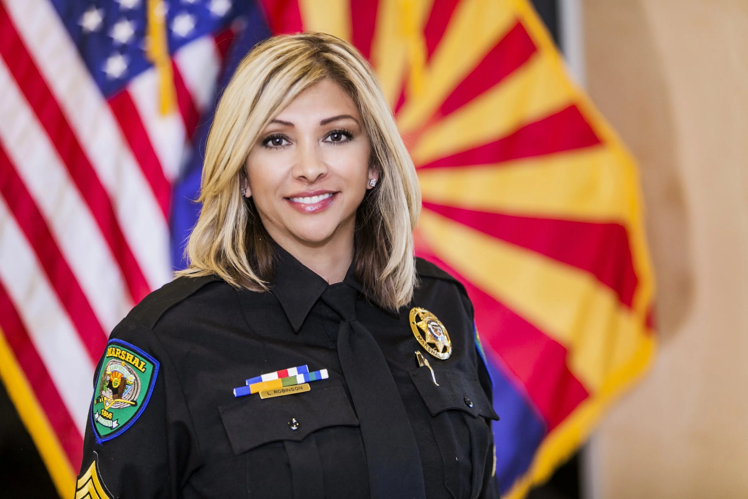A smiling female police officer in uniform, standing in front of American flags and another flag with red and yellow stripes.