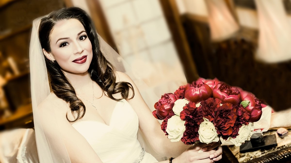 A bride with dark brown hair in curls, wearing a white dress with a sweetheart neckline, holding a bouquet of red and white flowers, with makeup including dark lipstick, standing indoors with warm lighting.