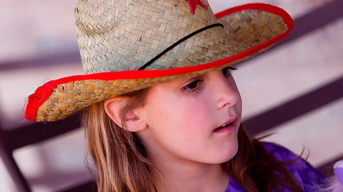 Young girl wearing a large straw hat with red and black accents, sitting outdoors with blurred background.