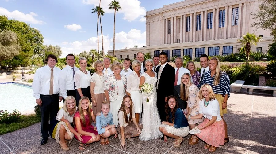 Group of people standing outdoors in front of a large building, celebrating a wedding with the bride and groom in the center.