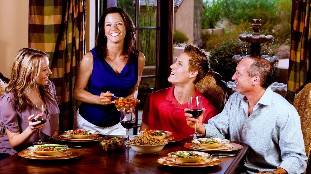 Family gathered around a dining table for a meal, with a woman serving food and the others smiling and holding glasses of wine.