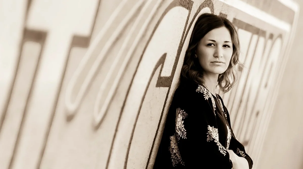 A woman standing against a wall with large painted letters, looking at the camera with a serious expression in sepia tone.
