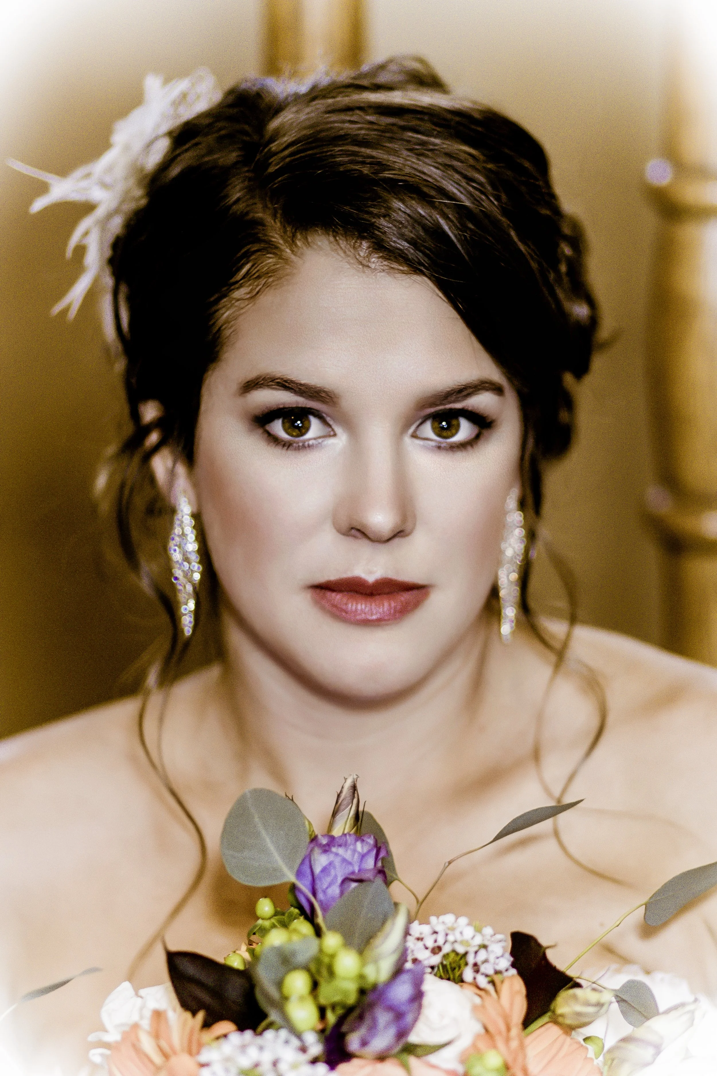 A woman with dark brown, shoulder-length hair, wearing chandelier earrings and holding a colorful bouquet of flowers, looking directly at the camera.