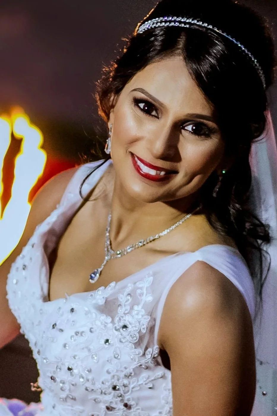 A woman in a white dress with embroidery and rhinestones, wearing a tiara and jewelry, smiling at the camera.