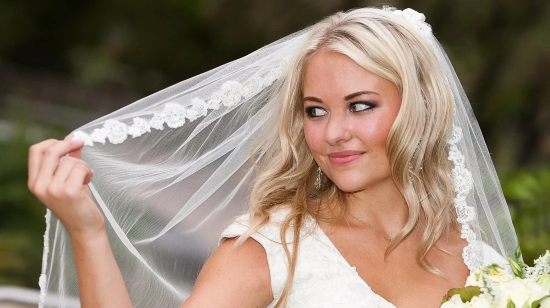 A bride with blonde hair, holding a veil with lace detail, outdoors on a wedding day.