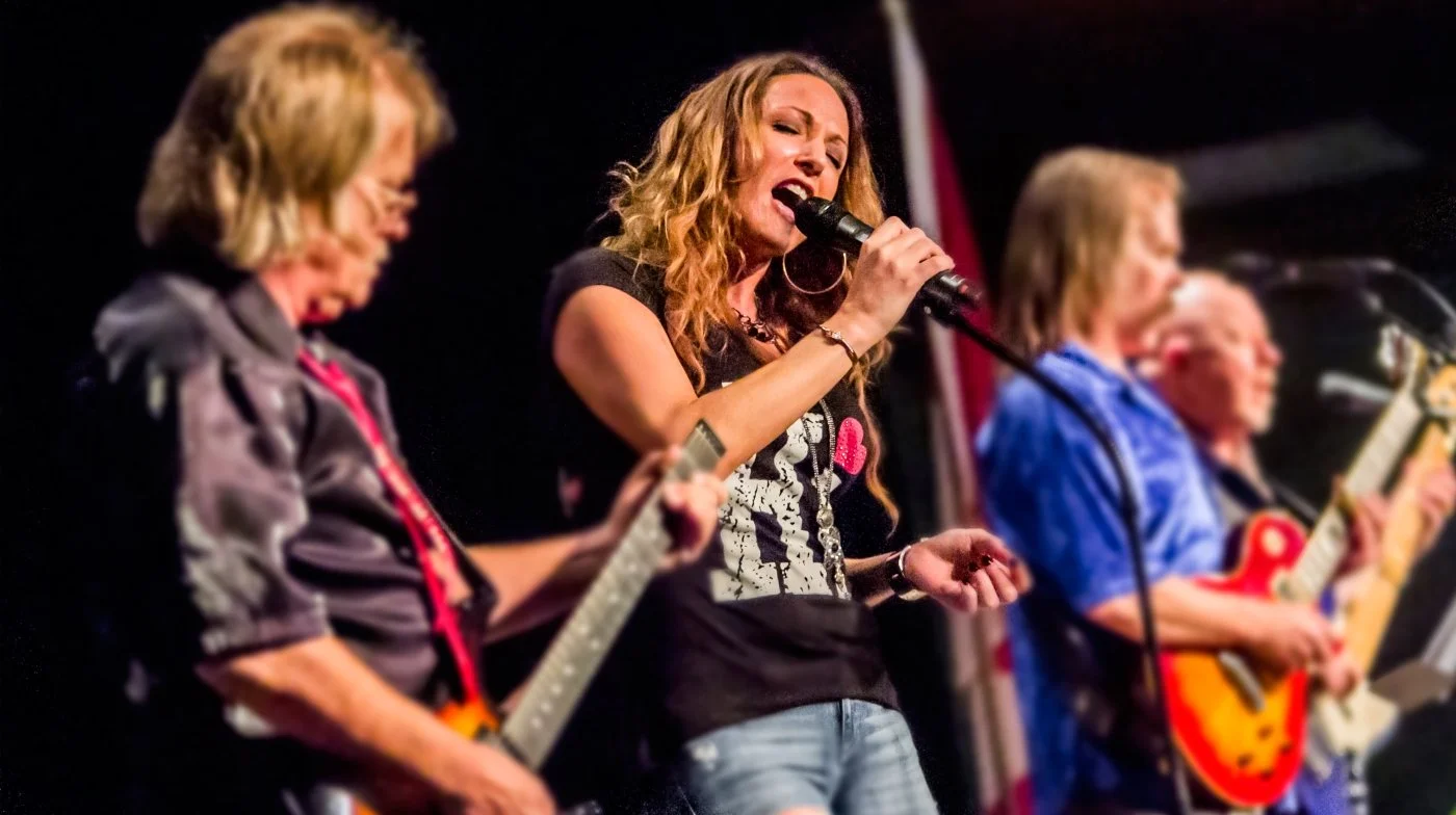 Four women performing on stage, three of them playing electric guitars and one singing into a microphone.