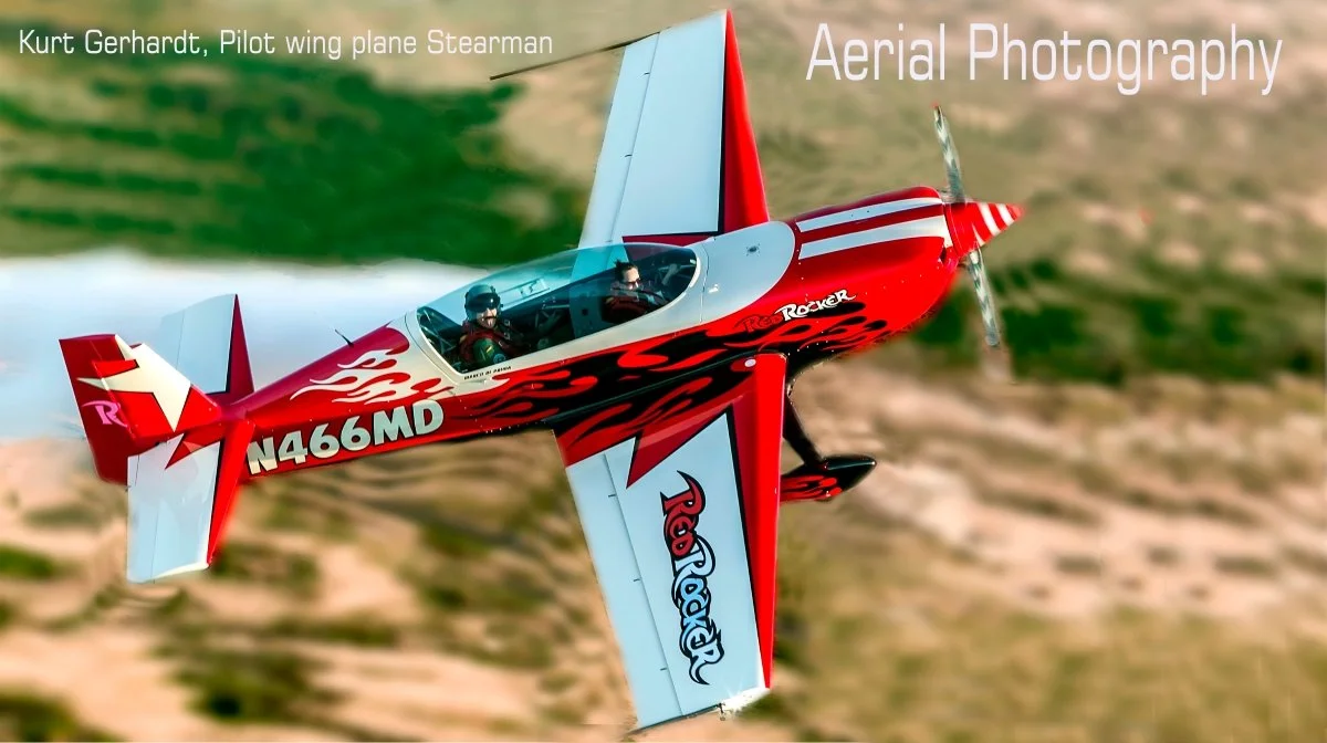 A red and white aerobatic airplane with flames and 'Red Roker' written on the wing, flying over grassy terrain with pilots visible inside.