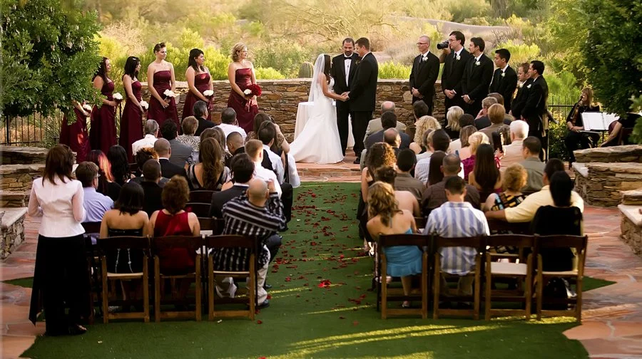 A wedding ceremony taking place outdoors with the bride and groom exchanging vows, surrounded by bridesmaids and groomsmen, with guests seated on wooden chairs, fallen rose petals on the grass path, and a scenic natural background.