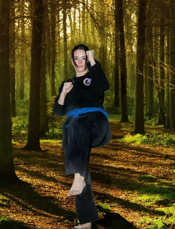 A woman practicing martial arts in a forest with sunlight streaming through trees.