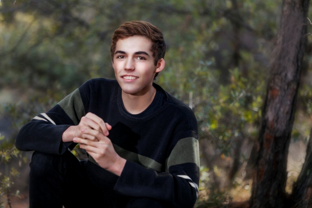 Young man with brown hair and a friendly expression, sitting outdoors in a wooded area, wearing a black sweater with green and white stripes, with trees and greenery in the background.