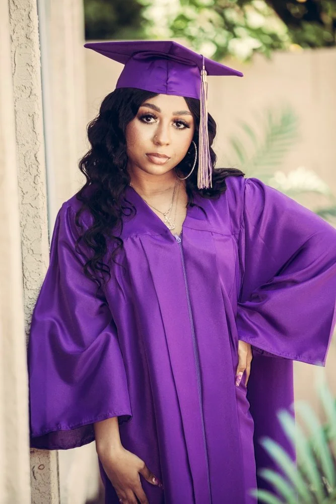 A young woman in a purple graduation gown and cap, standing outdoors with greenery in the background.