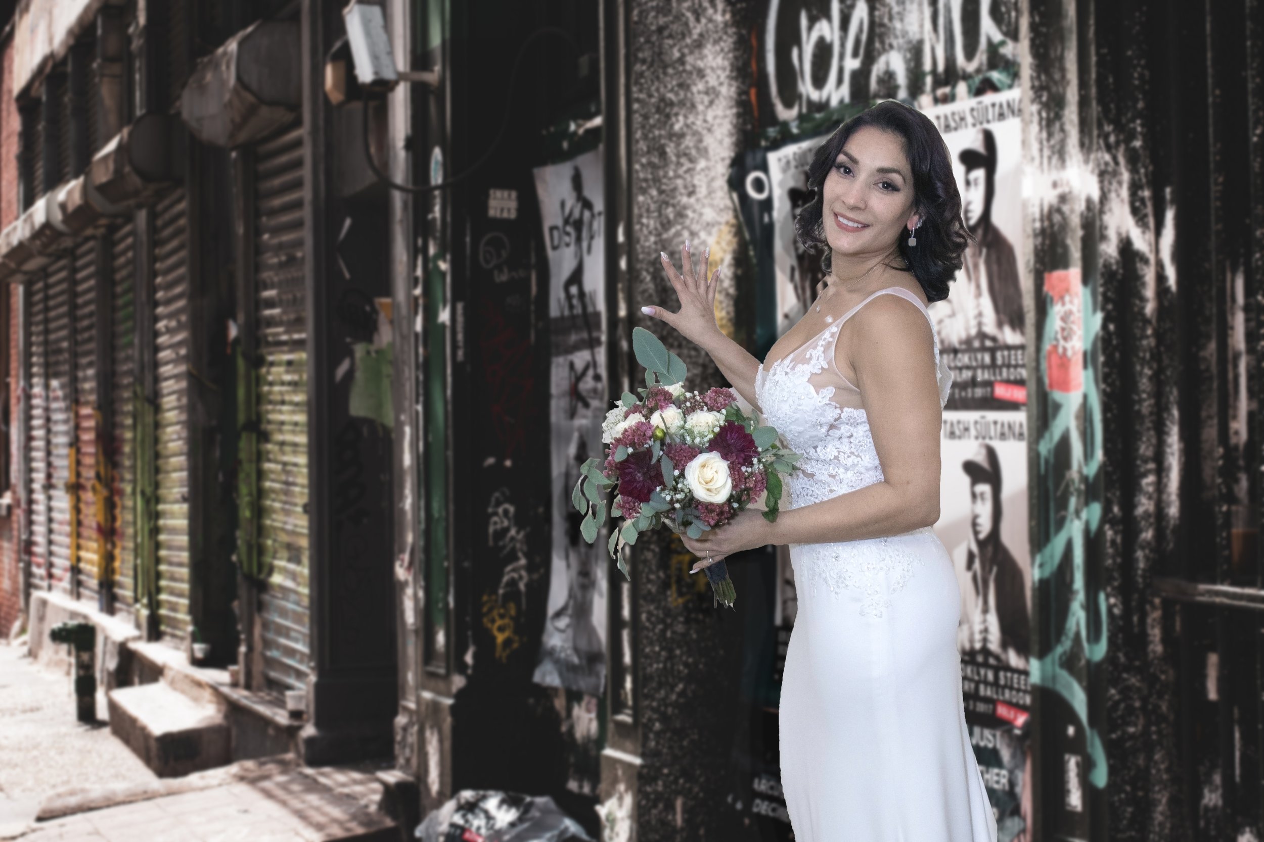 A woman in a white wedding dress holding a bouquet of flowers, standing in front of a graffitied wall with posters and graffiti art.