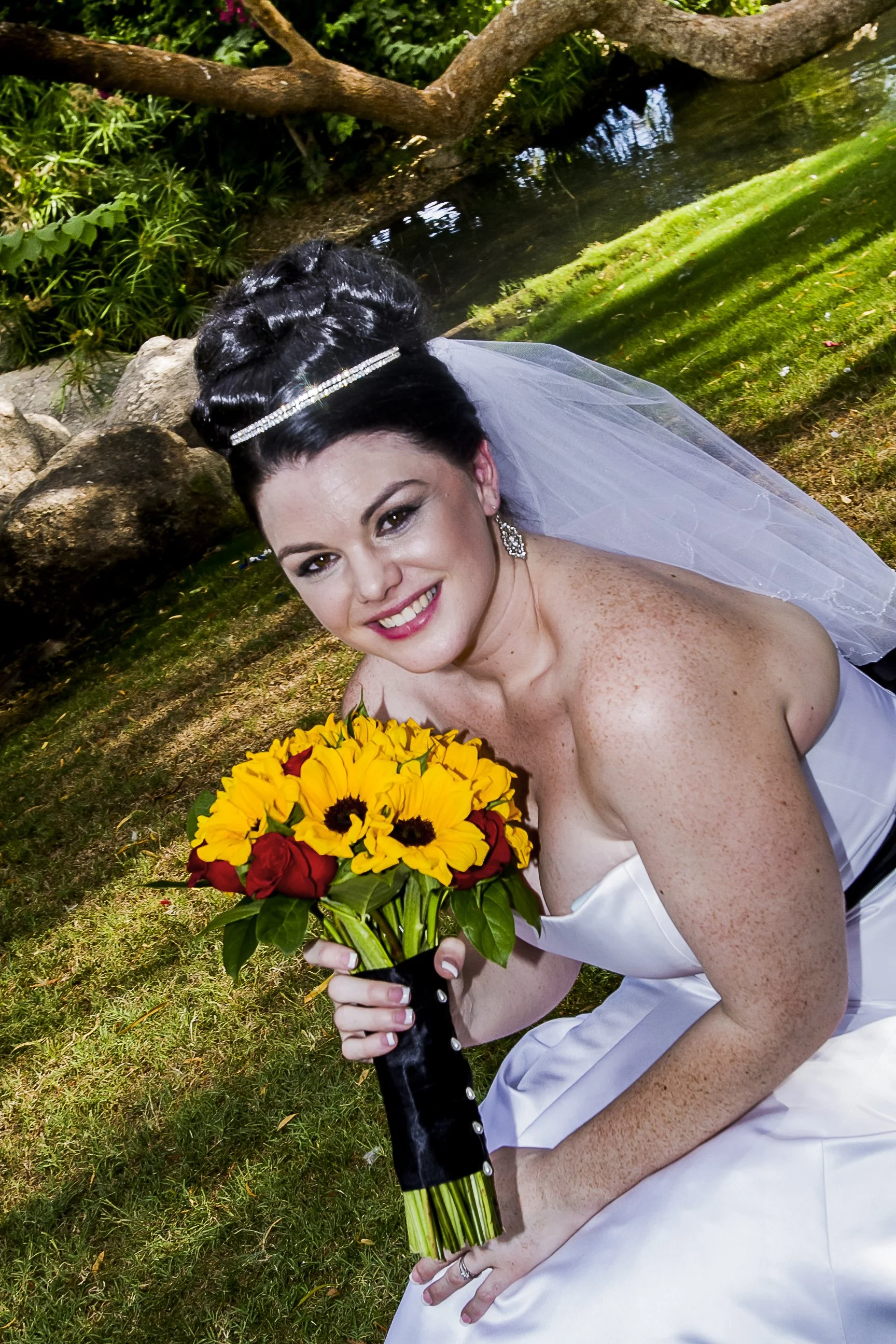 A smiling bride in a white wedding dress holding a bouquet of yellow and red roses and sunflowers, sitting outdoors near a pond with lush green foliage and rocks.
