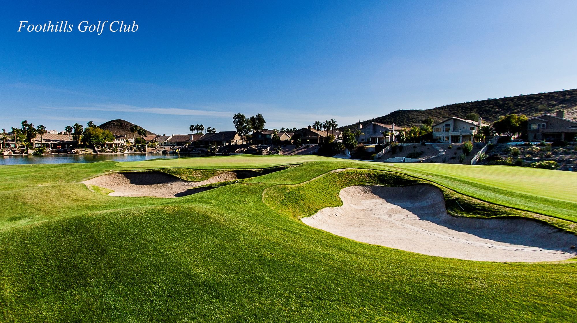 Golf course with sand bunkers, green grass, water hazard, and houses in the background under a blue sky.