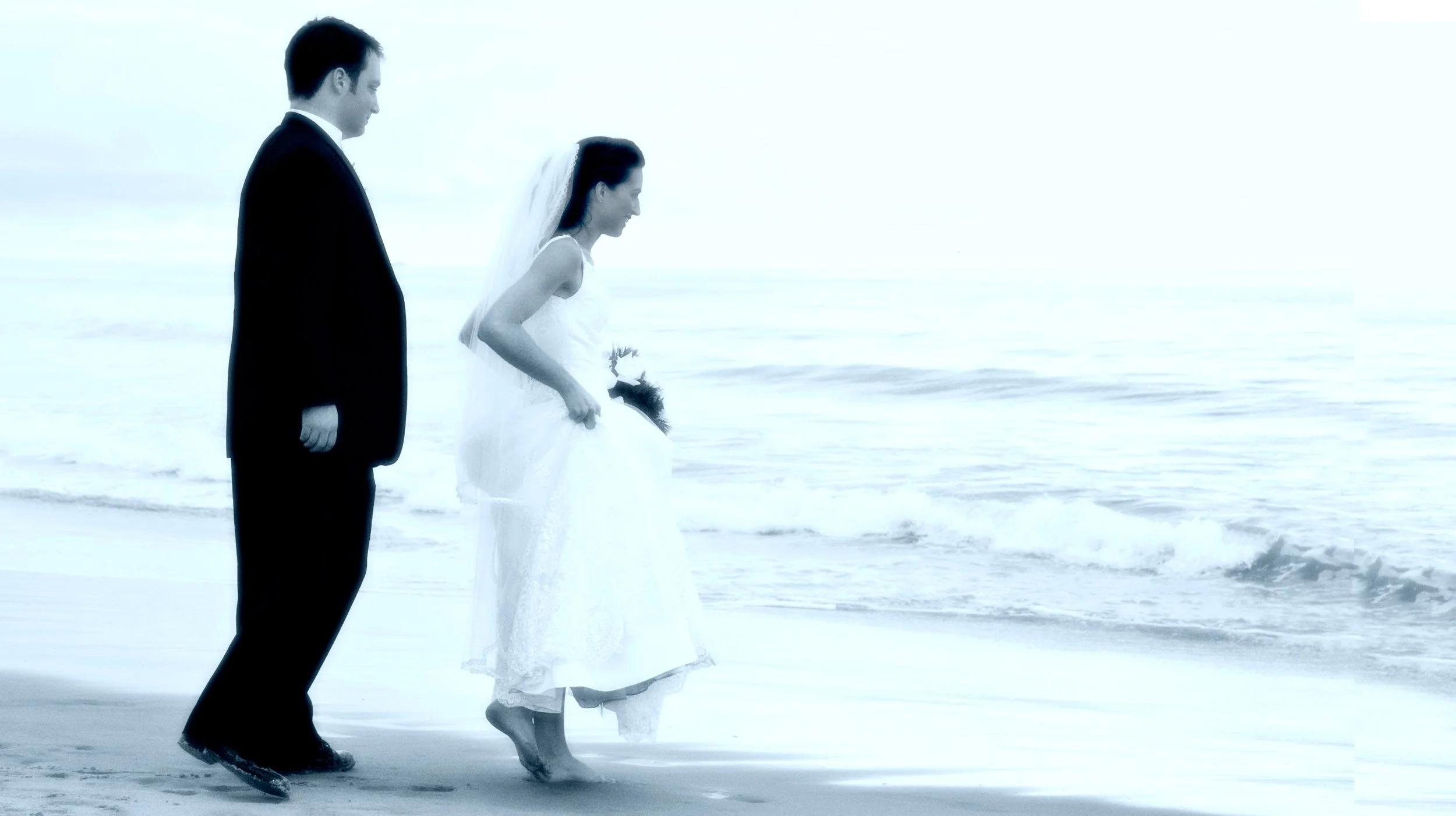 A bride and groom walking barefoot on the beach near the water while holding hands, dressed in wedding attire.