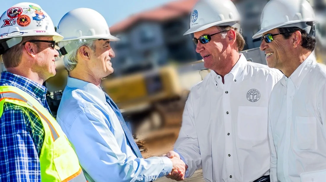 Four men in safety helmets and white shirts, two shaking hands at a construction site, with heavy machinery and buildings in the background.