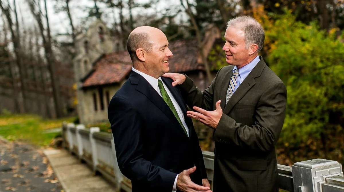 Two middle-aged men in suits talking and smiling outdoors near a bridge with a scenic background of trees and a building.