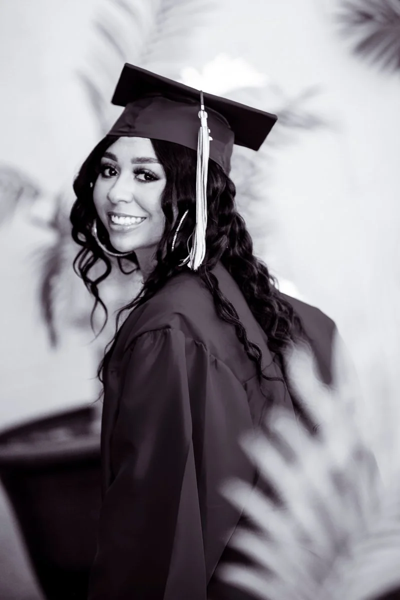 Black-and-white photo of a young woman in a graduation cap and gown, smiling and looking over her shoulder.