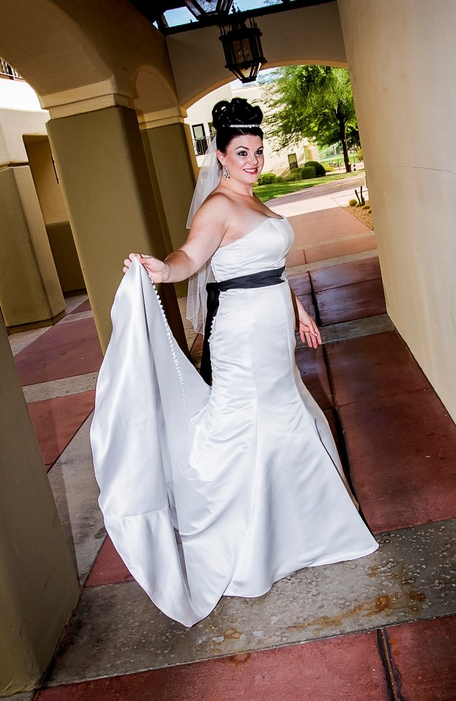 Woman in a strapless white wedding dress with a black sash, holding the train of her gown, standing under an archway outdoors.