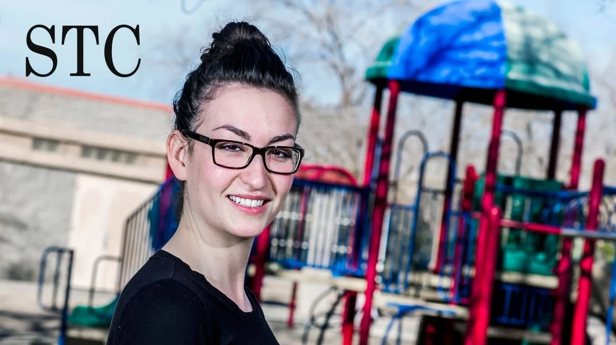 Smiling woman in glasses standing outdoors near a colorful playground.