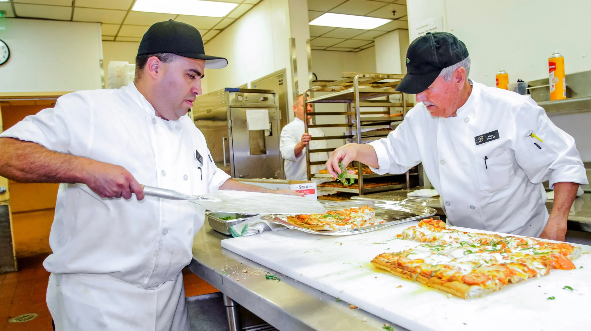Two chefs in white uniforms and black hats prepare pizza in a commercial kitchen. One chef is slicing the pizza, while the other is adding garnishes. In the background, another worker can be seen holding a tray.