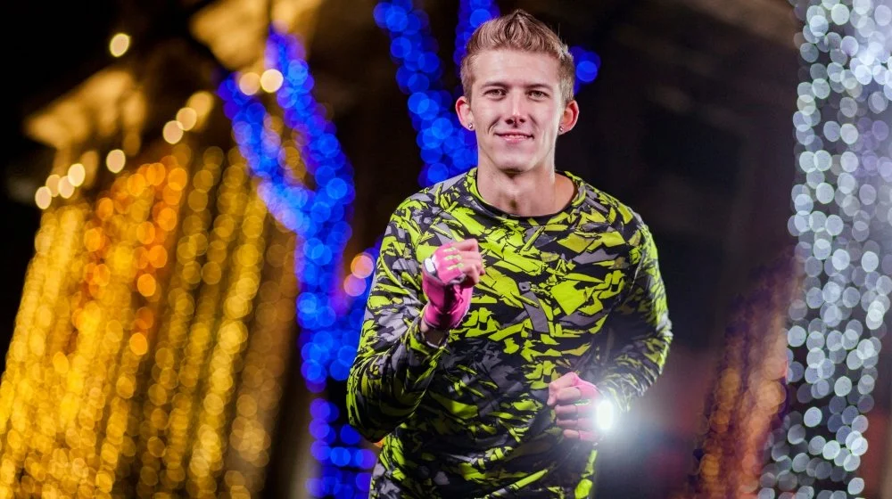 A young man with light skin and short light hair jogging outdoors at night, wearing a neon green and black patterned long-sleeve shirt and pink gloves, with colorful blurred holiday lights in the background.