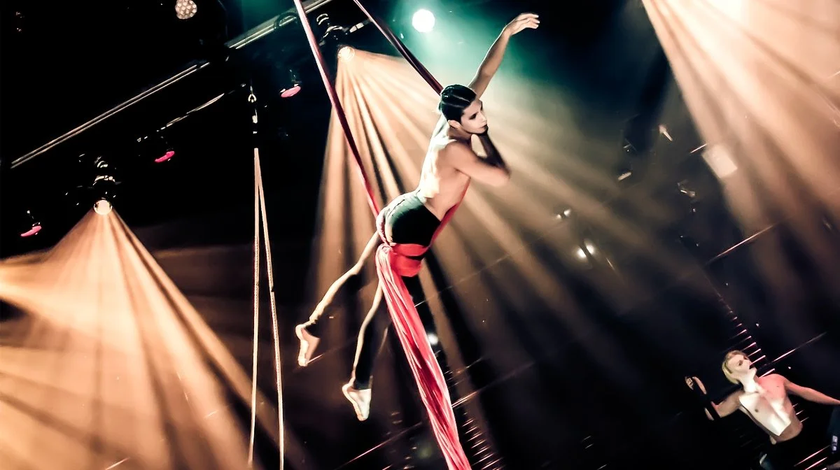 Two aerial performers hanging on silks in a circus or acrobatic performance, with dramatic lighting and dark background.