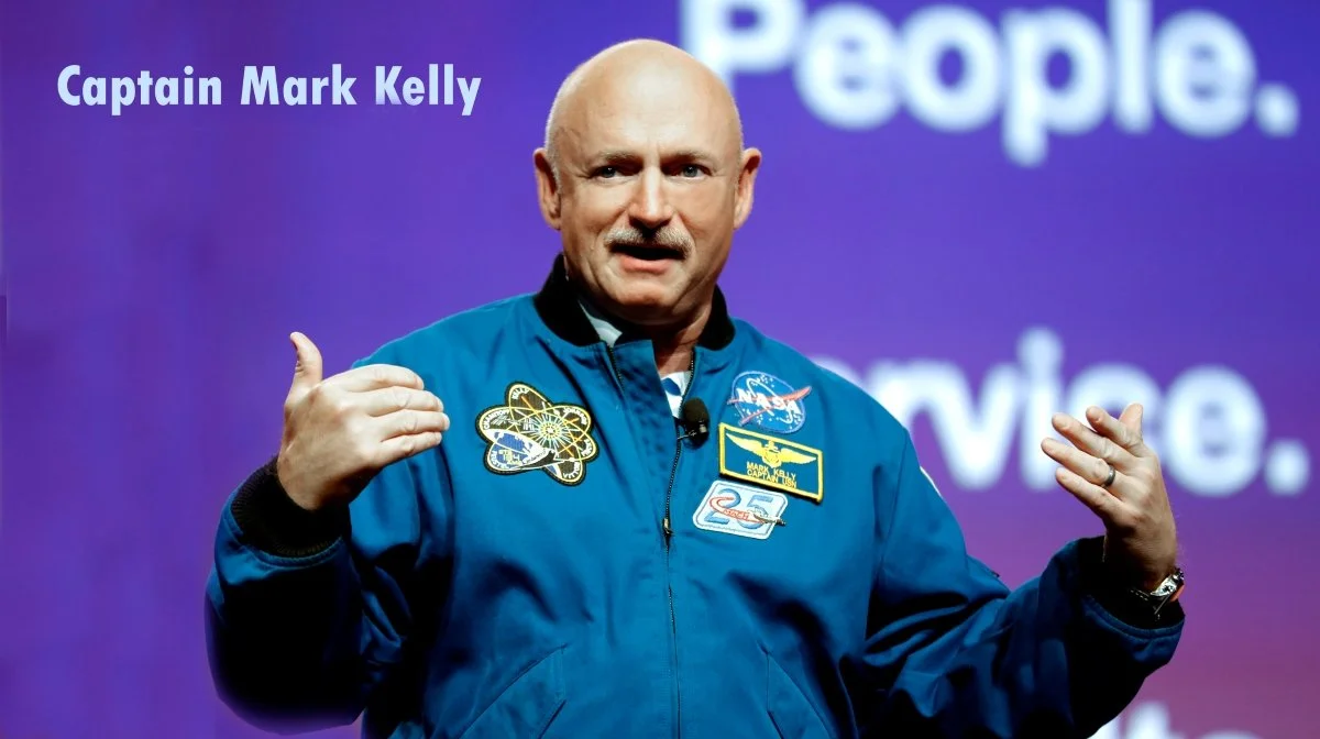 Captain Mark Kelly in a blue NASA jacket giving a presentation with his hands raised, with a purple background displaying the words "People" and “Trust” in white.
