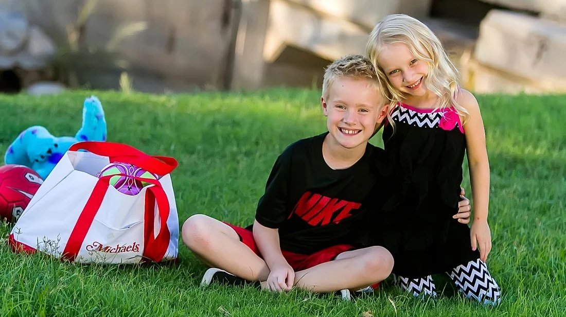 A boy and girl sitting on grass at a park, smiling. The boy is wearing a black Nike T-shirt and red shorts, the girl is wearing a black dress with chevron patterns and a pink bow. There is a Michaels shopping bag and plush toys nearby.