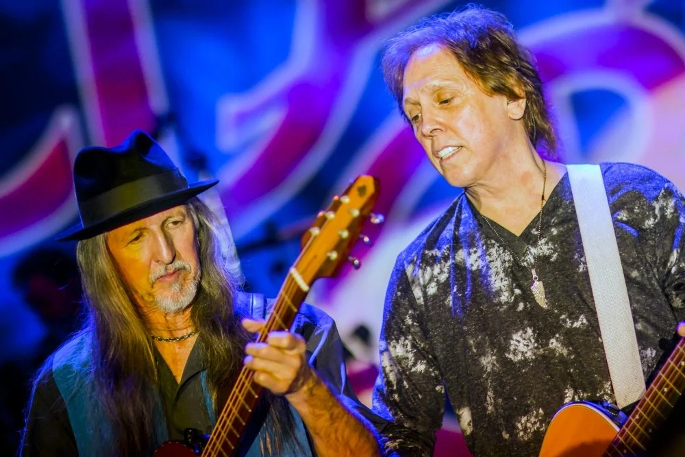 Two men playing guitars together, one wearing a black hat and long hair, the other with short hair and a patterned shirt, in a colorful indoor setting.