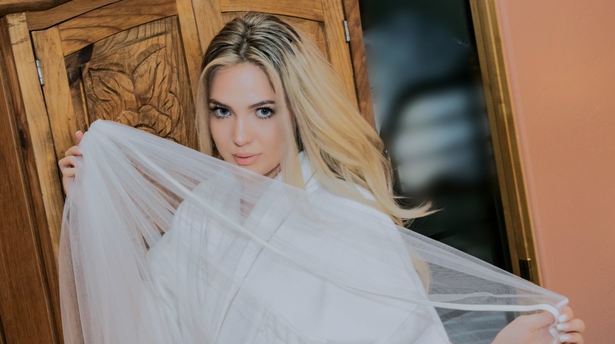 A young woman with long blonde hair and blue eyes holds a sheer white veil in front of her face, standing in front of a wooden door.