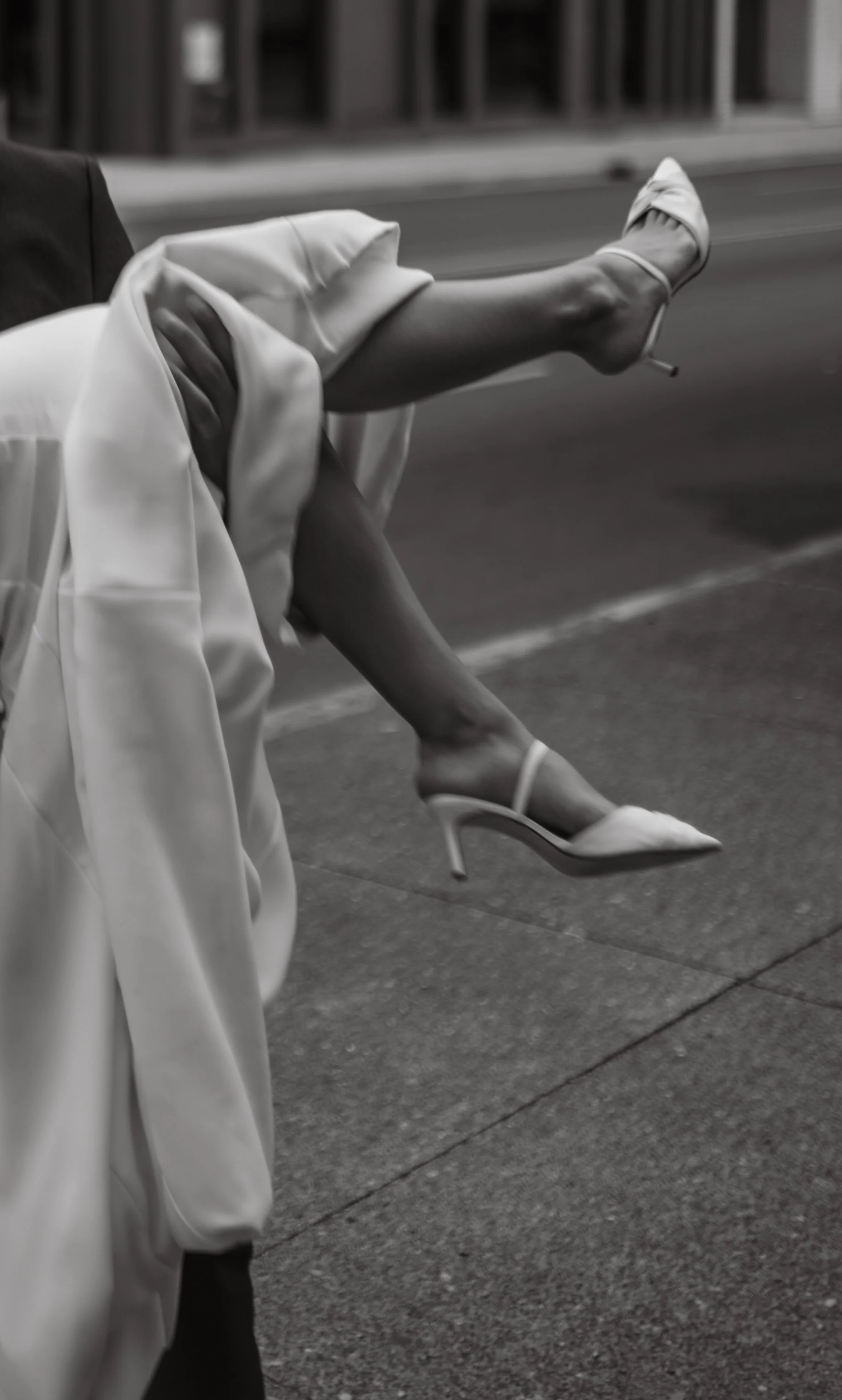Black and white photo of a person carrying another person wearing a dress and heels on a street.