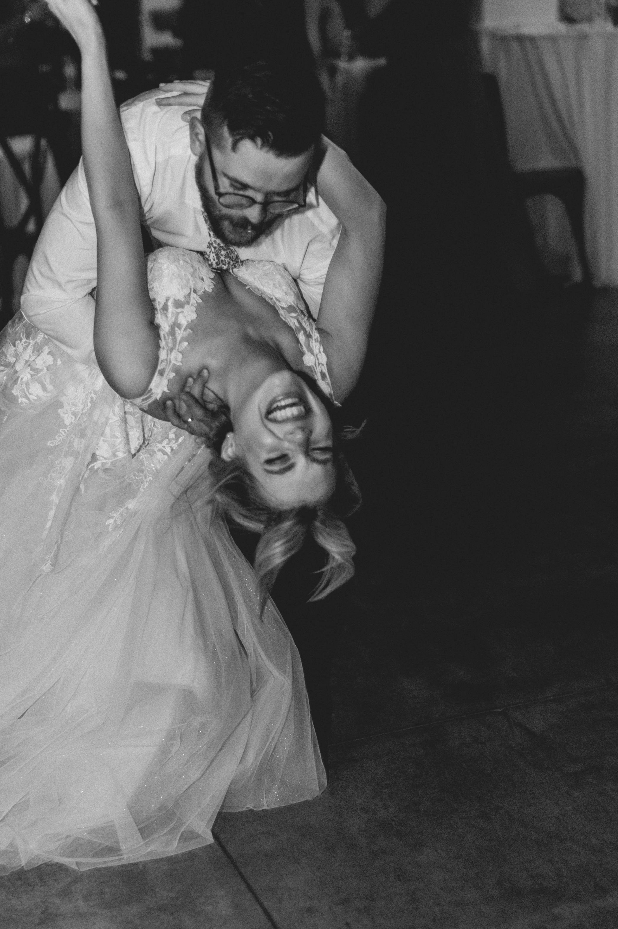 A black-and-white photo of a couple dancing, with the man dipping the woman backwards. The woman in a wedding dress is laughing with joy.
