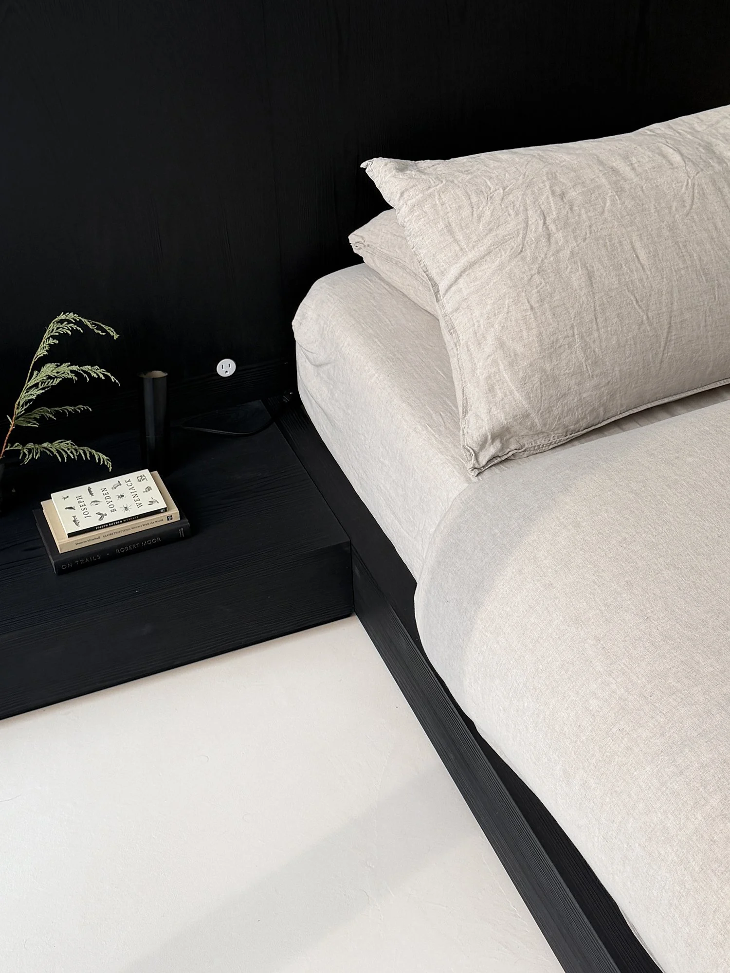 Low-profile blackened oak platform bed and matching minimalist nightstand styled with natural linen sheets and books.