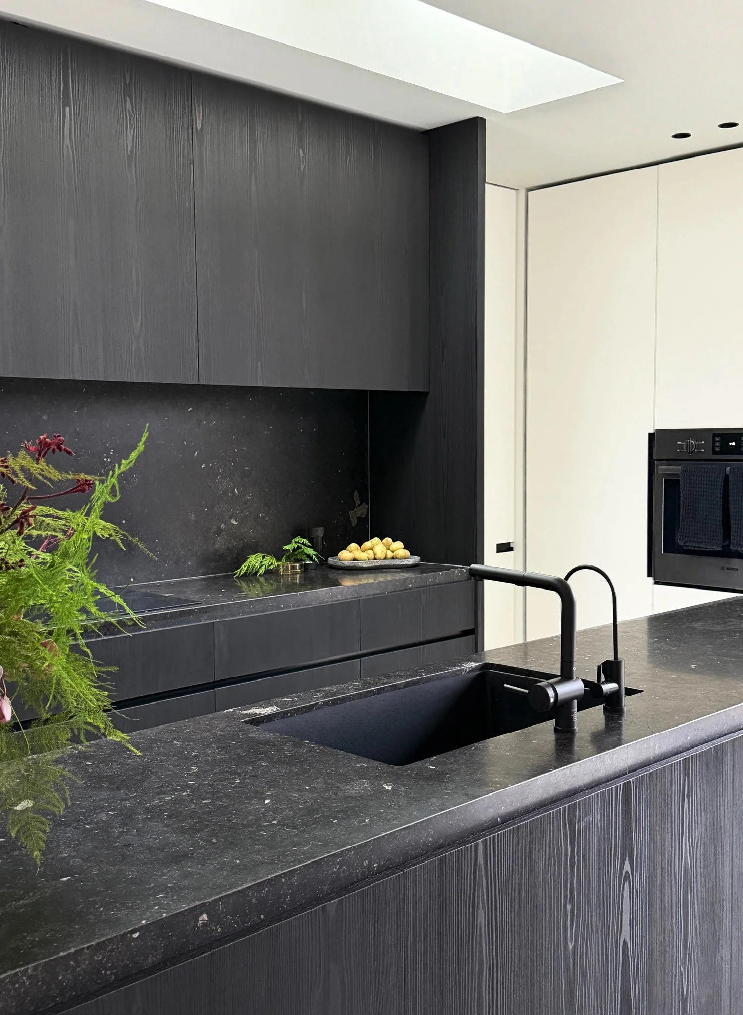Soft brutalist kitchen design featuring blackened oak cabinetry, continuous dark stone countertops and backsplash, and a matte black faucet.