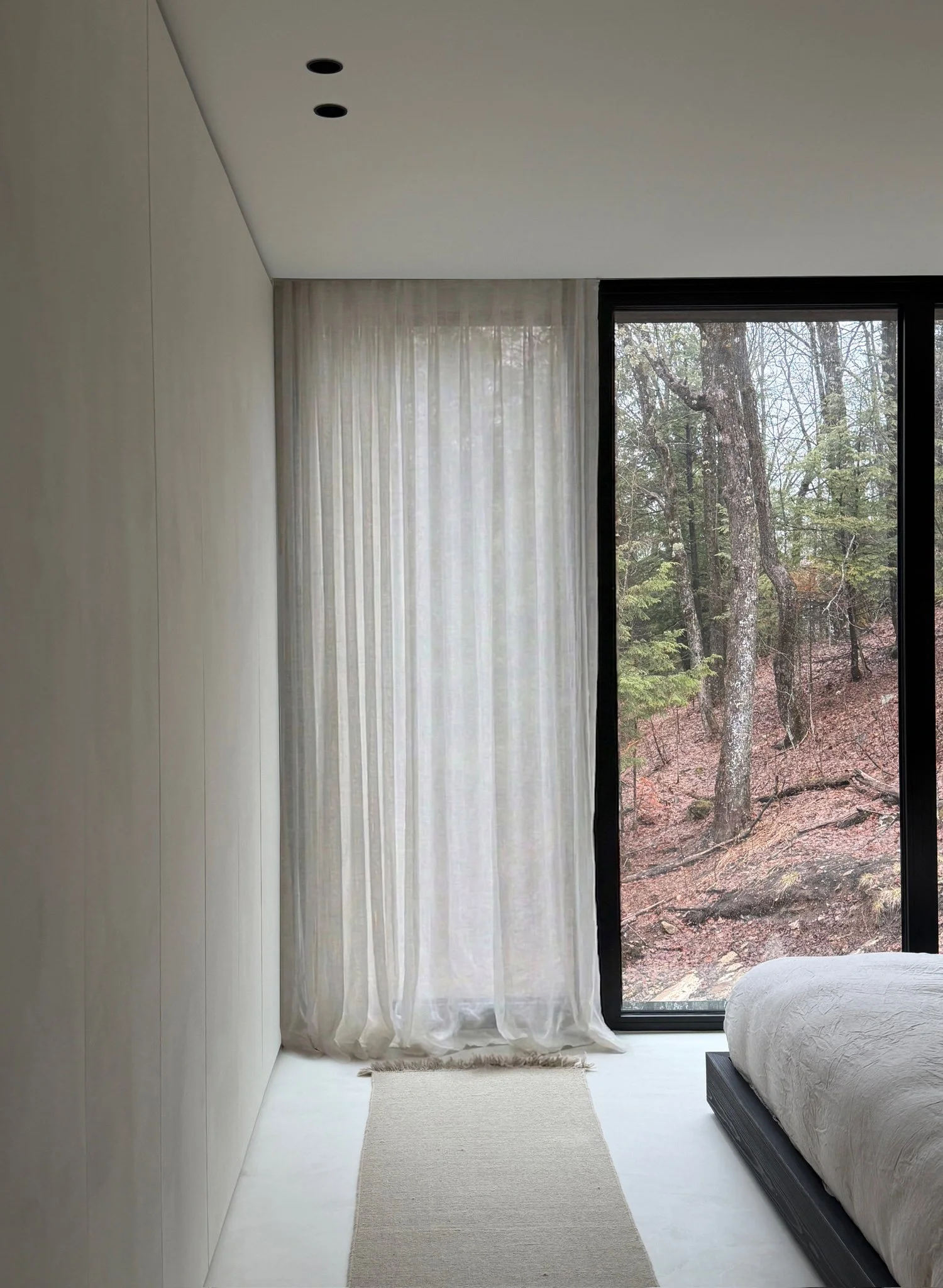 Serene minimalist bedroom corner featuring smooth plaster walls, sheer linen curtains, and a black-framed window overlooking a wooded landscape.