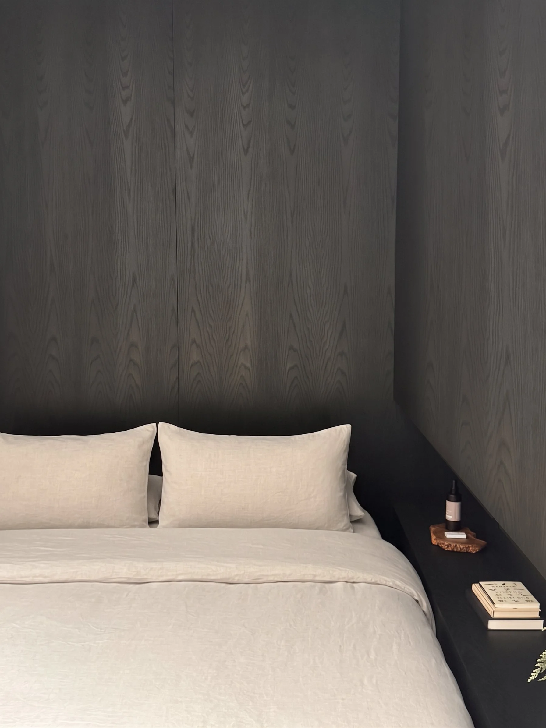 Soft brutalist bedroom detail with a dark wood wall, natural linen pillows, and a built-in floating nightstand with greenery.