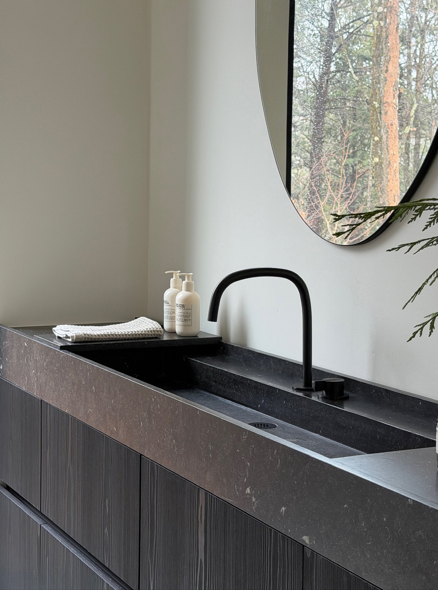 Custom dark stone bathroom trough sink and ebonized oak vanity paired with a matte black faucet and minimalist round mirror.