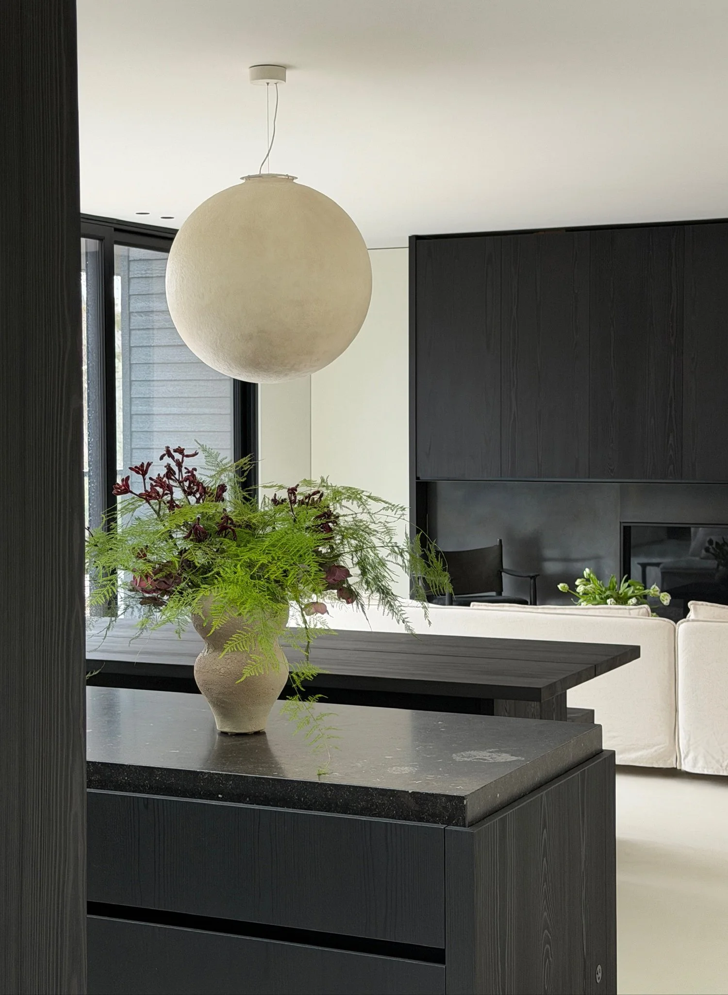 Open concept soft brutalist space featuring an ebonized oak dining table, dark stone island, and a large sculptural plaster globe pendant.