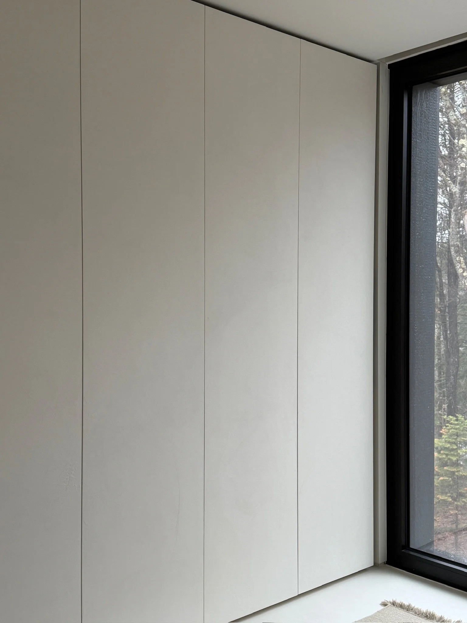 Floor-to-ceiling seamless plaster wardrobe doors beside a large black-framed window in a clean, minimalist interior.