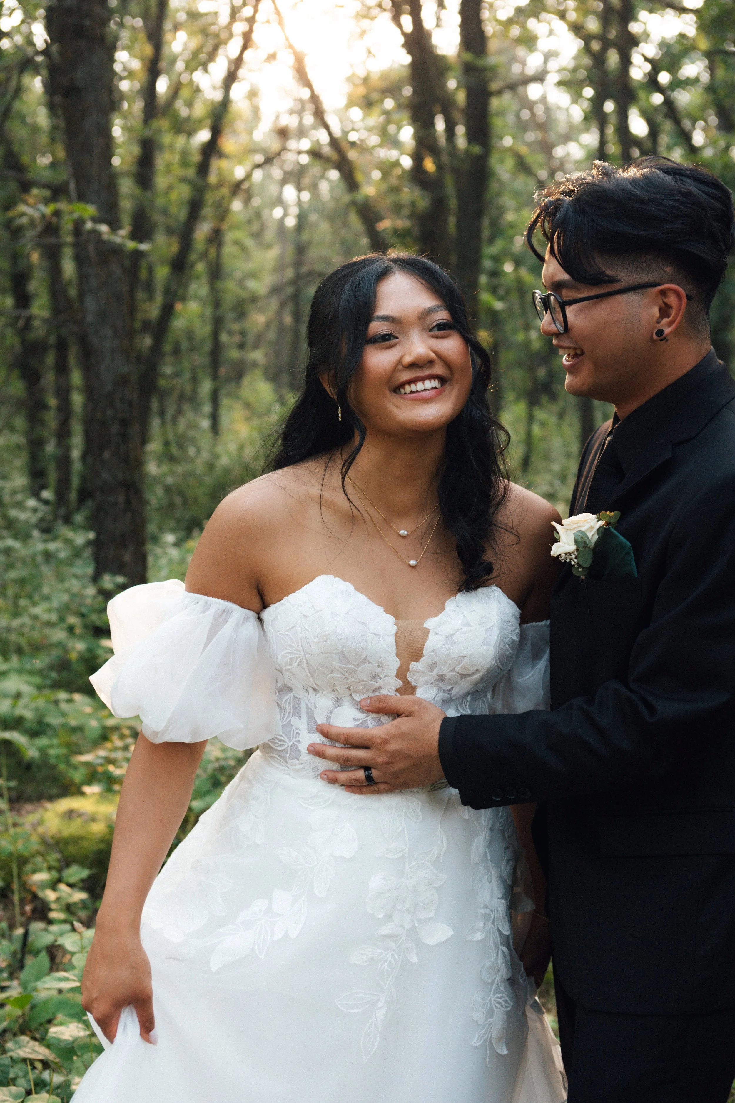 A newlywed couple smiling and facing each other in a forest setting during their wedding, with the bride in a white off-shoulder gown and the groom in a black suit with a boutonniere.