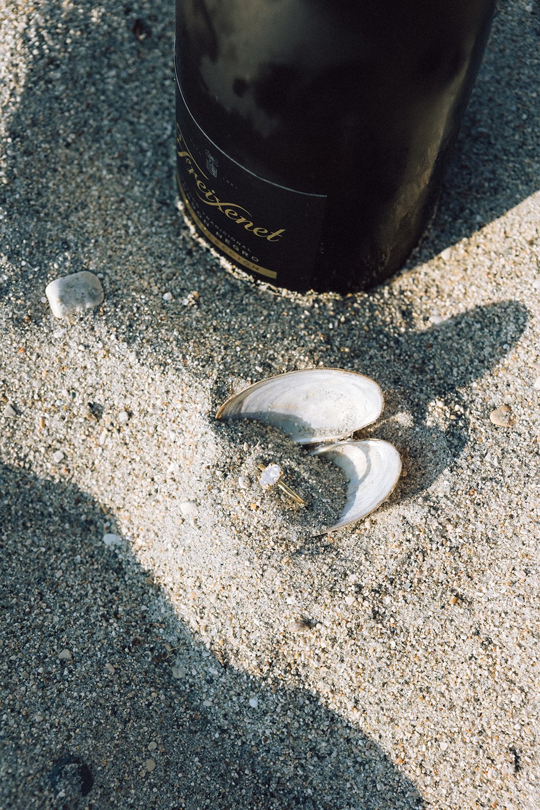 A wine bottle, seashells, and a ring are on sandy ground at the beach.