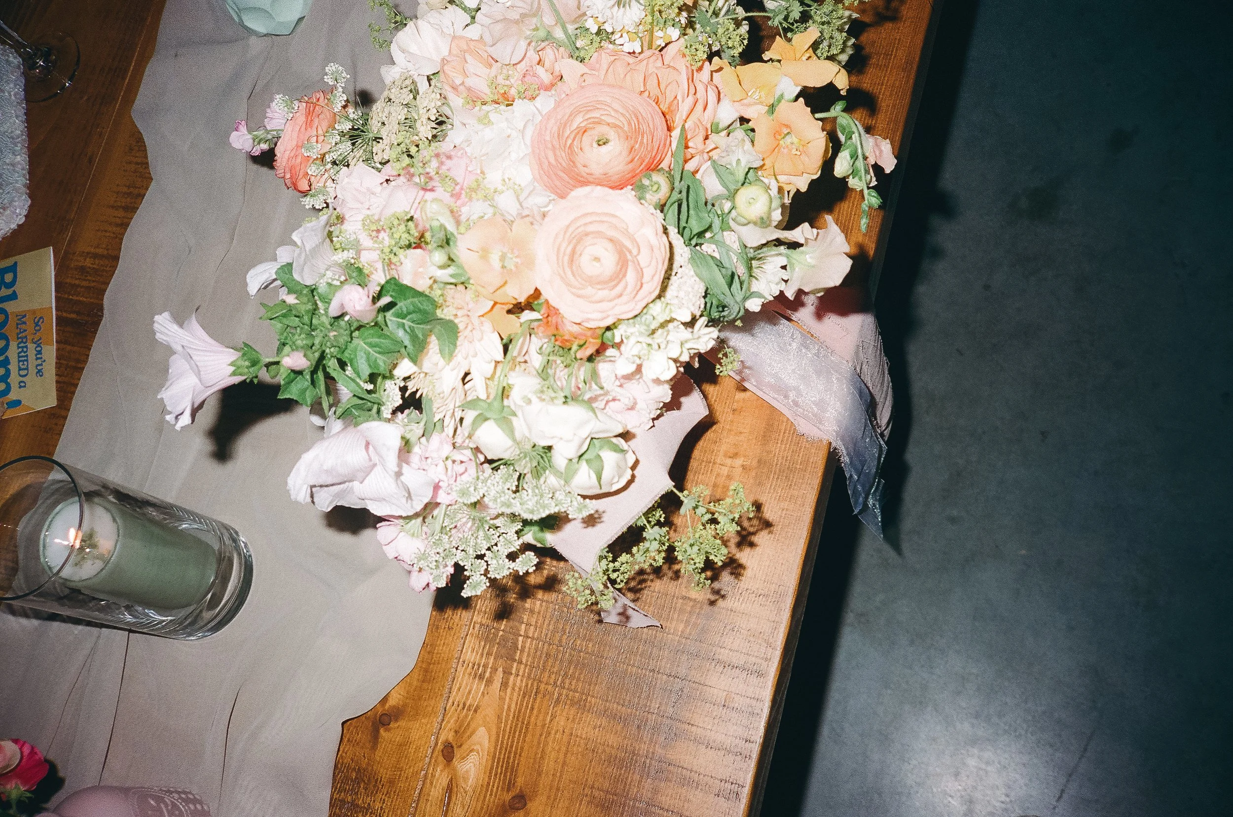 A floral arrangement of pink and white flowers with green foliage on a wooden table, accompanied by a candle in a glass holder and a piece of gray fabric underneath.