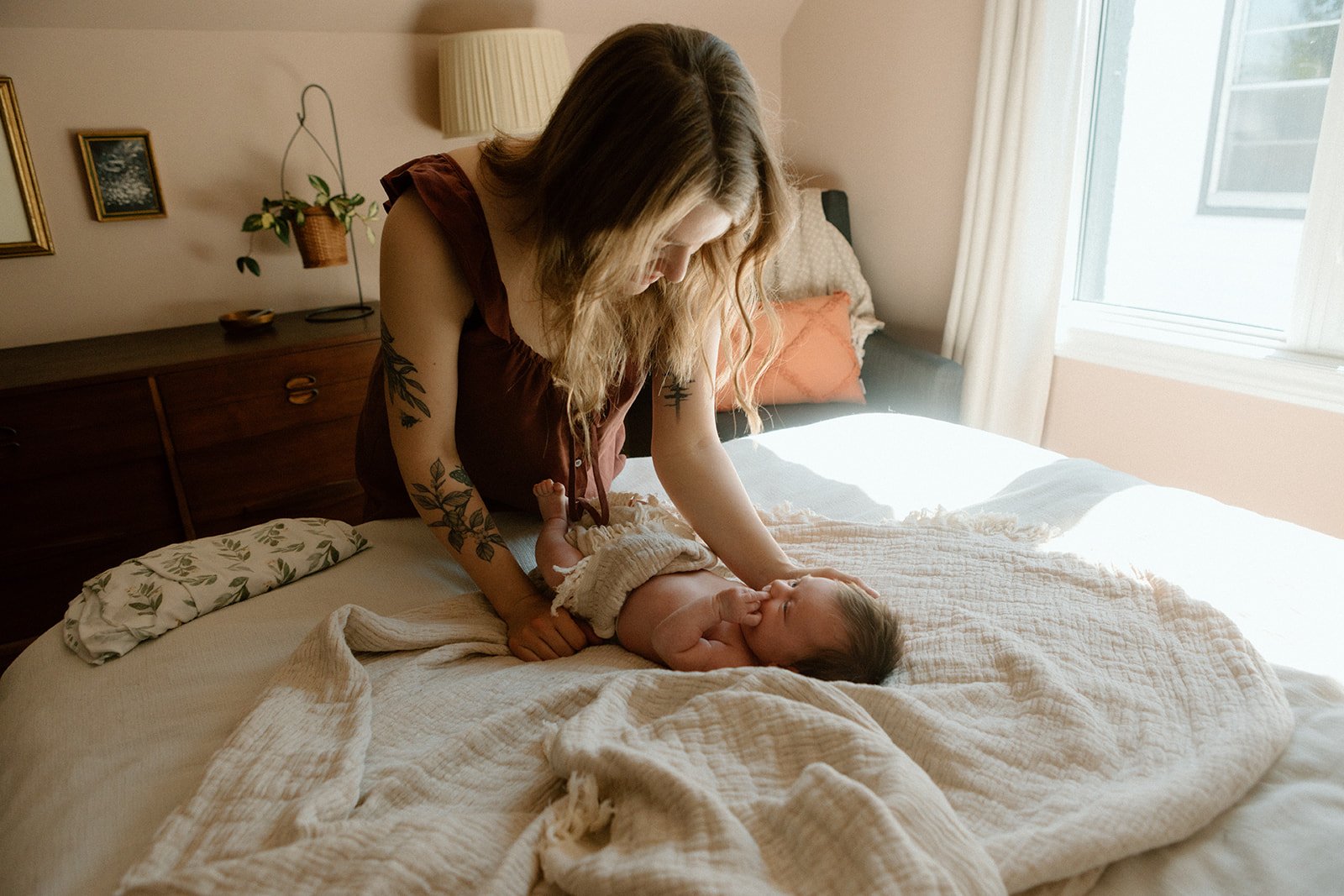 A woman with tattoos and loose blonde hair is leaning over a baby lying on a bed, gently touching the baby's face in a sunlit bedroom.