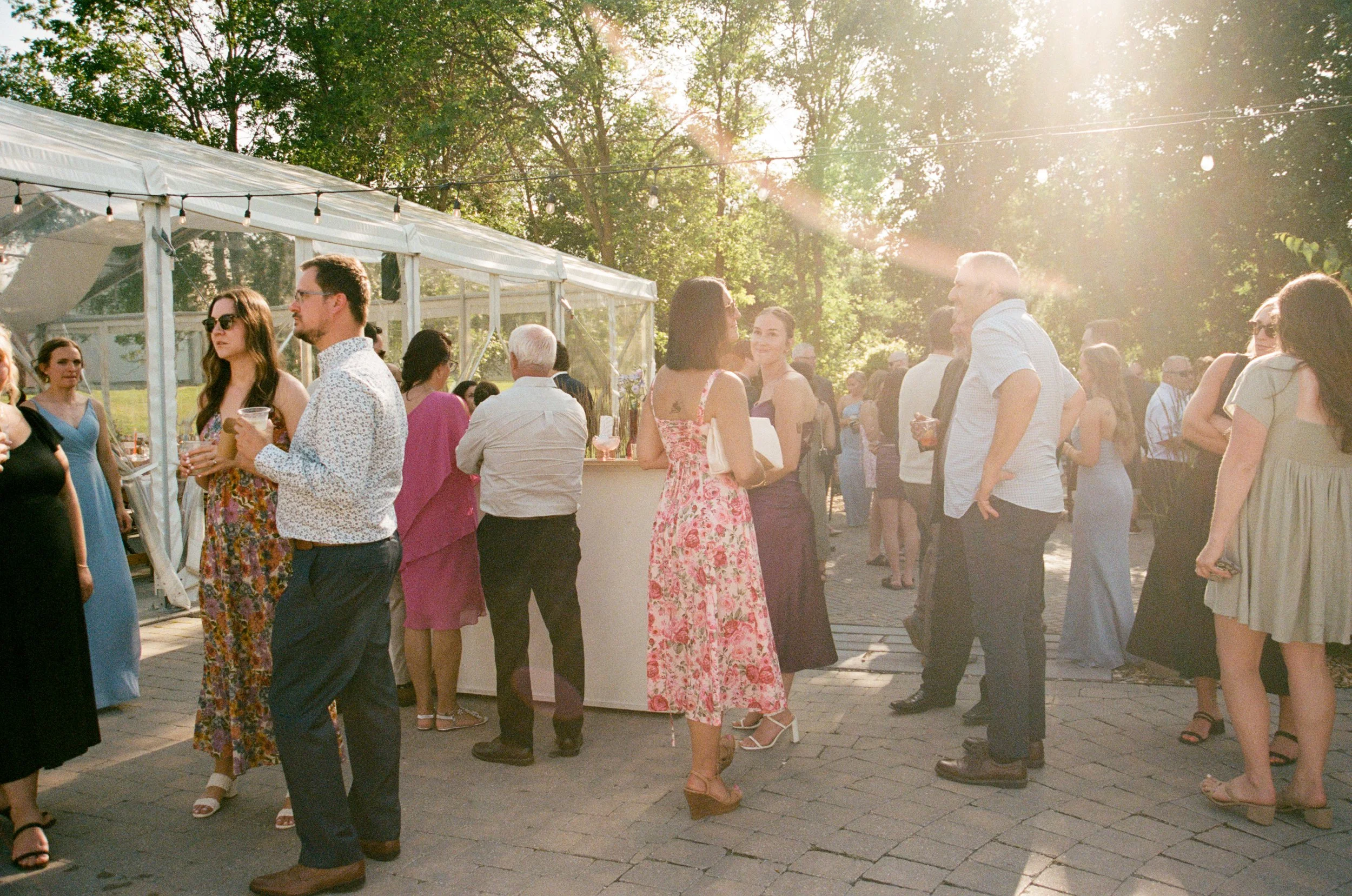 People socializing at an outdoor event during sunset, with a white tent and trees in the background.