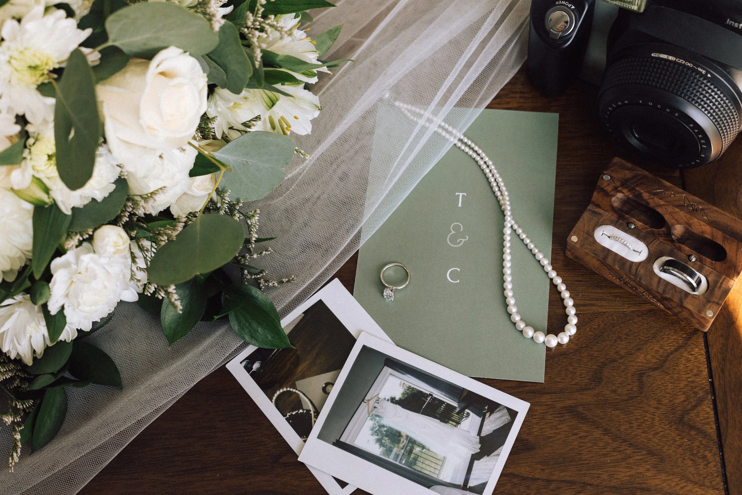 Arrangement of wedding items including white flowers, greenery, a white tulle fabric, a pearl necklace, a ring with a diamond, a green card with initials 'T & C', two instant photographs, a camera, and wedding bands in a wooden box.