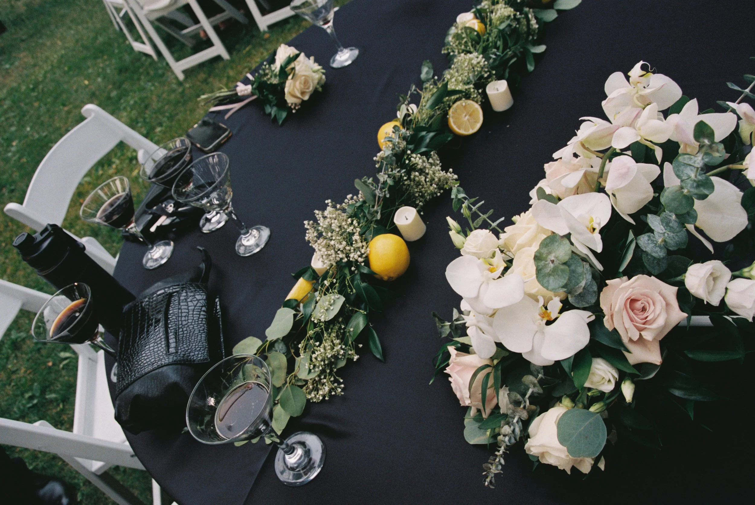 Elegant outdoor dining table decorated with white and blush flowers, greenery, candles, lemon slices, and lemon fruits on a black tablecloth, set with wine glasses and black dinnerware.