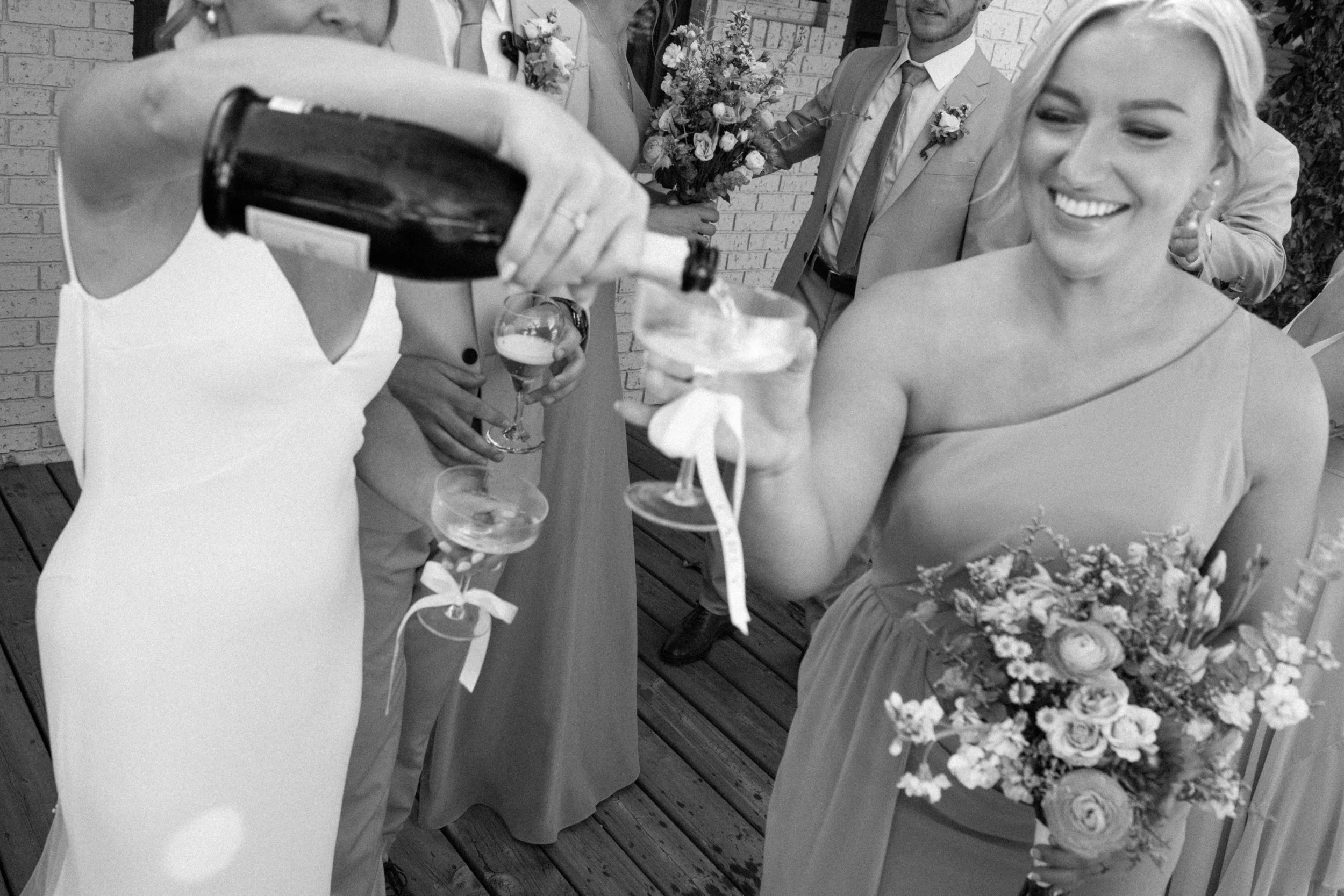 Women toasting with drinks at a wedding reception, with a focus on a smiling woman holding a bouquet, and a person pouring champagne.