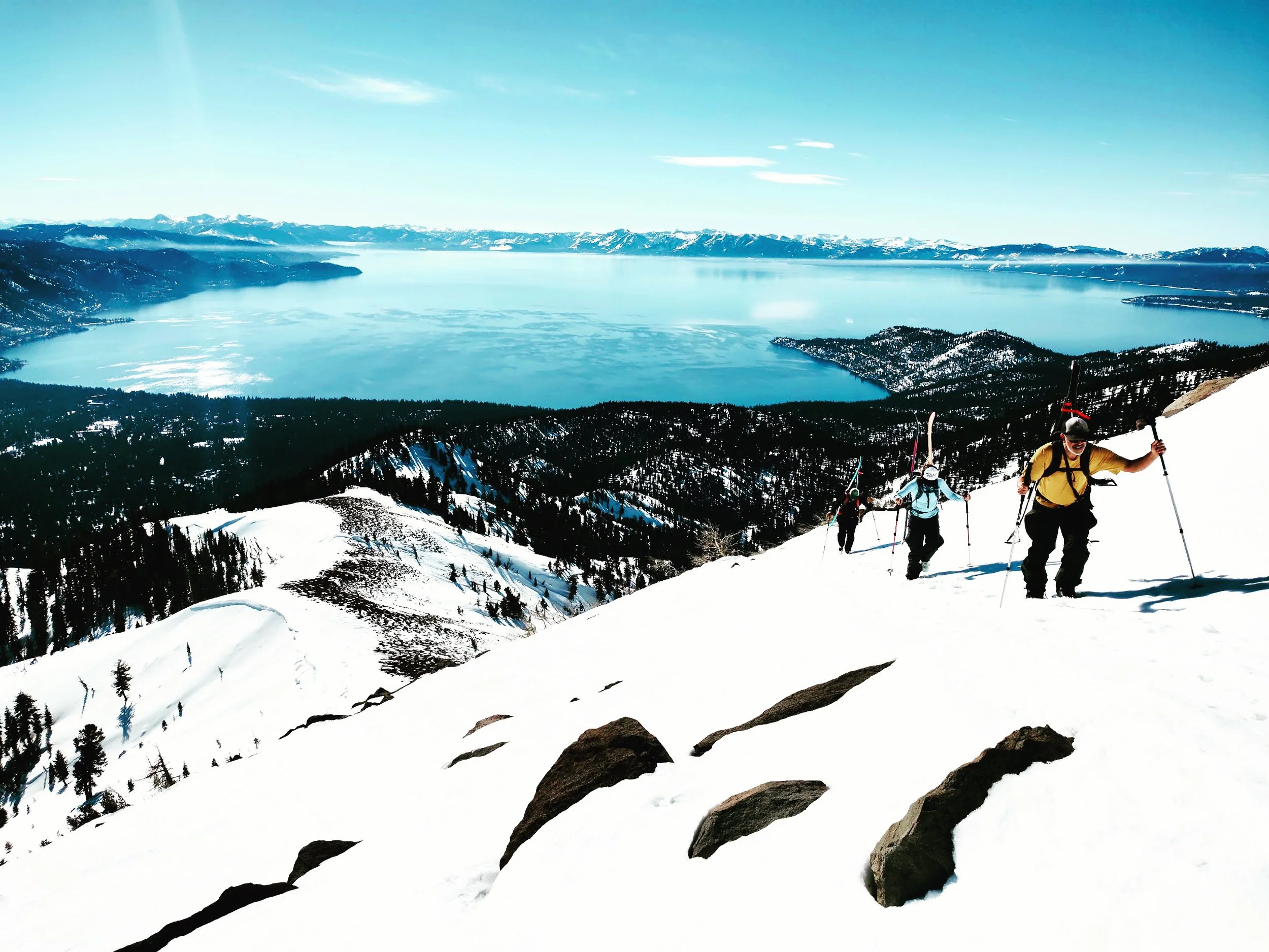 Three splitboarders hike up a snow covered slope above Lake Tahoe on a spring day.