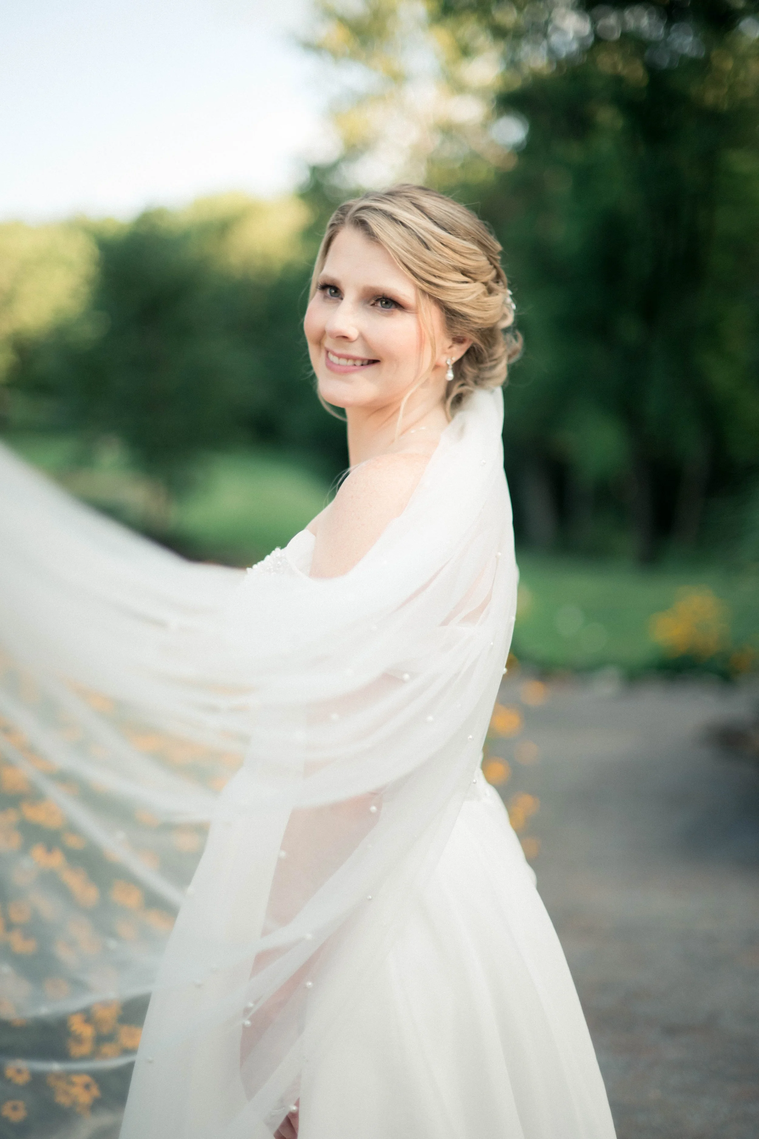 A smiling bride in a white wedding dress outdoors, with a sheer veil flowing, and trees in the background.